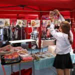 A woman organizes art prints in a vibrant outdoor market stall with colorful clothing and accessories.